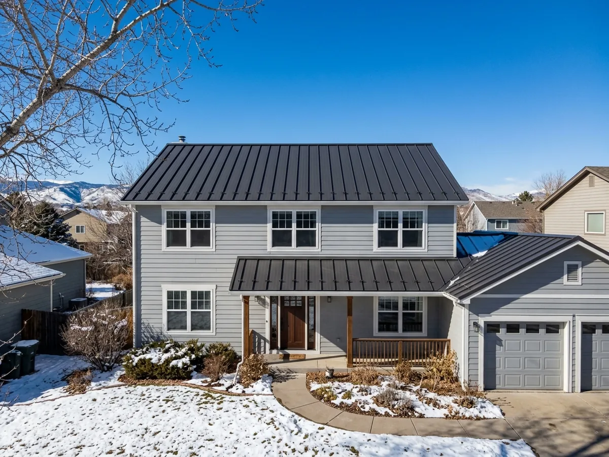 Gray two-storey house with a dark standing seam metal roof over the main house, porch and garage, light snow on the lawn and snowy mountains behind